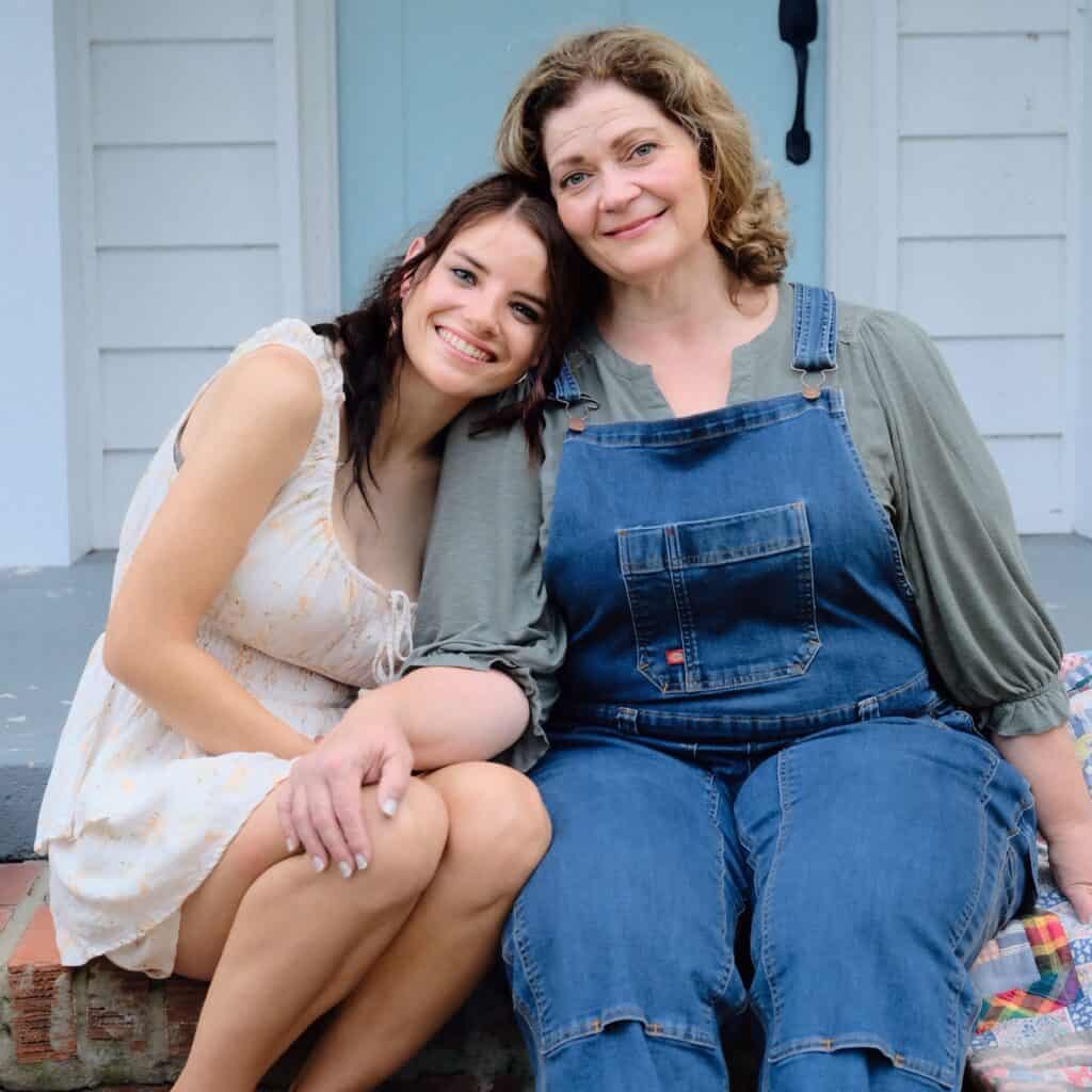 Two women sit close together on a porch step, smiling. One wears a light dress, the other blue overalls and a green shirt. The woman in the dress leans her head on the other’s shoulder affectionately.