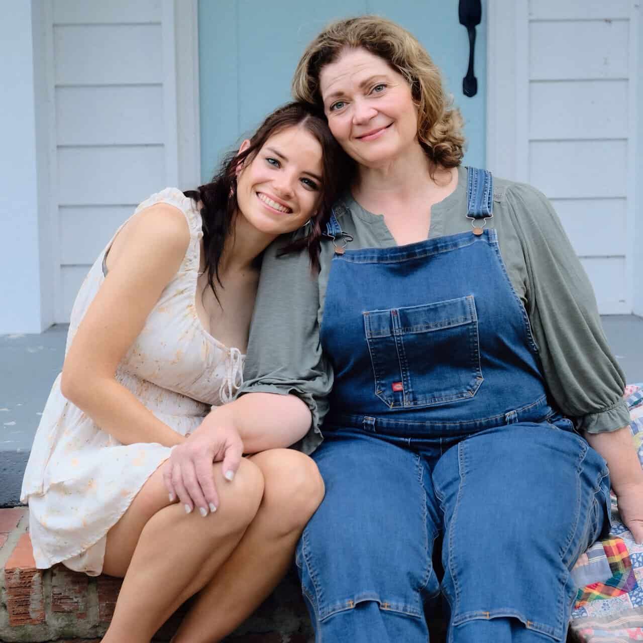 Two women sit close together on a porch step, smiling. One wears a light dress, the other blue overalls and a green shirt. The woman in the dress leans her head on the other’s shoulder affectionately.