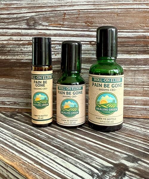 Three bottles of "Pain Be Gone Topical Oil" from Healing Green are shown on rustic wooden planks; the differently sized bottles have tan labels with green and gold accents.