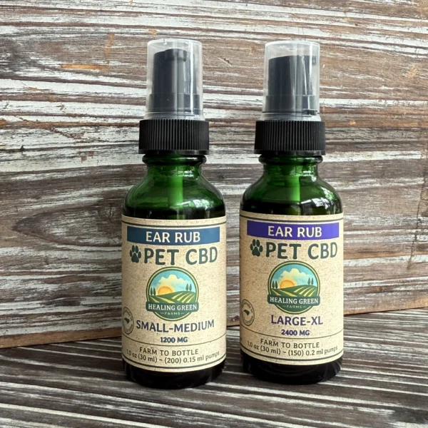 Two green glass bottles of Healing Green Ear Rub pet CBD with pump tops sit on a wooden surface. One is labeled "Small-Medium" (1200 mg) and the other "Large-XL" (2400 mg).
