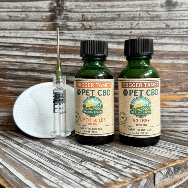 Two bottles of Trigger Tamer, a syringe, and a small textured pad are arranged on rustic wood. The bottles are labeled for pets up to 50 lbs, each containing 1200 mg CBD.