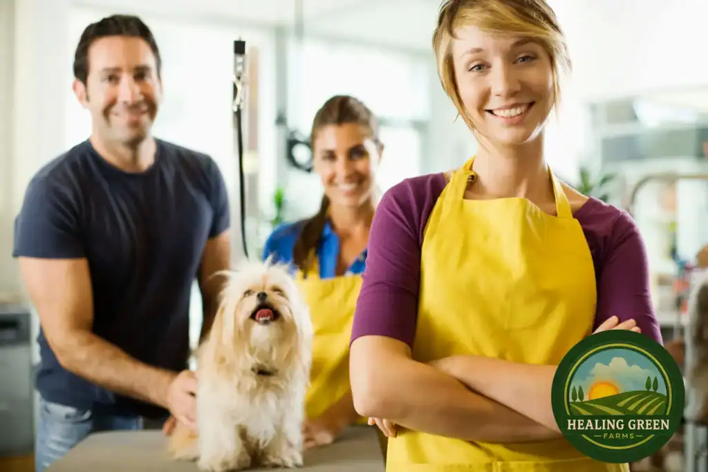 Three smiling people in yellow aprons stand with a fluffy white dog on a table in a bright room. A "Healing Green Farms" logo appears in the lower right corner of the image.