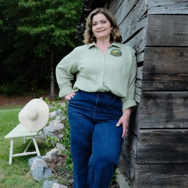 A woman in a light green shirt and blue jeans leans against a rustic wooden building outdoors, smiling. A white hat rests on a chair nearby, with green trees and rocks in the background.