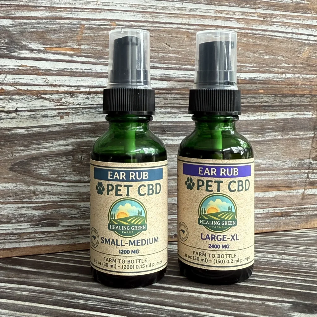 Two green glass bottles of Healing Green Ear Rub pet CBD with pump tops sit on a wooden surface. One is labeled "Small-Medium" (1200 mg) and the other "Large-XL" (2400 mg).